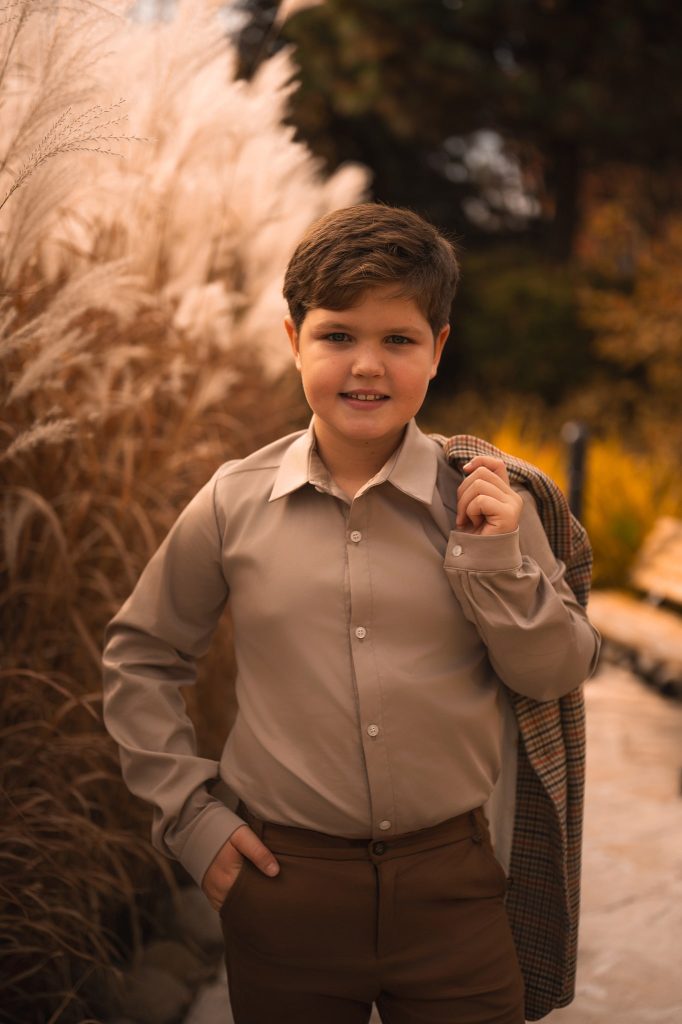 Beżowa koszula chłopięca prezentowana w eleganckiej jesiennej stylizacji – klasyczny fason z kołnierzykiem, idealna na komunię, uroczystości rodzinne lub święta. Ujęcie na tle traw ozdobnych podkreśla ciepłą, stonowaną kolorystykę i ponadczasowy charakter ubrania