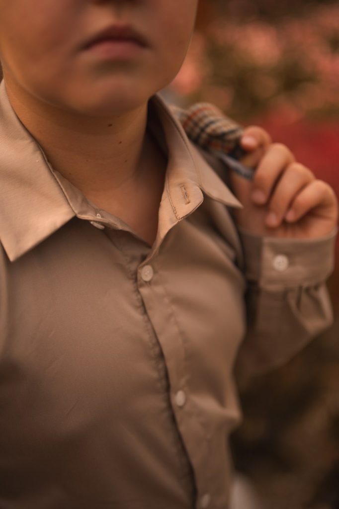 Beżowa koszula chłopięca prezentowana w eleganckiej jesiennej stylizacji – klasyczny fason z kołnierzykiem, idealna na komunię, uroczystości rodzinne lub święta. Ujęcie na tle traw ozdobnych podkreśla ciepłą, stonowaną kolorystykę i ponadczasowy charakter ubrania